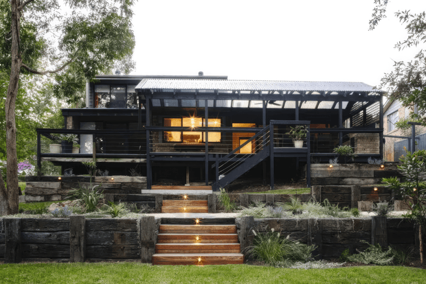 Exterior view of a raised, dark-clad contemporary house set within a landscaped sloping garden. The home features broad timber-framed glazing, covered outdoor verandah or balcony areas, a corrugated metal roof, black balustrades, and a central timber stair descending through tiered timber retaining walls. Warm interior lighting contrasts with the dark exterior finishes.
