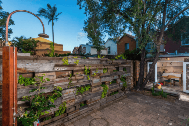 Outdoor courtyard or patio area with a rustic reclaimed-timber vertical garden wall, climbing plants, brick paving, mature trees, and a distinctive copper-toned curved outdoor shower fixture. A glazed opening at right reveals an indoor dining or living area, with neighbouring residential buildings beyond.