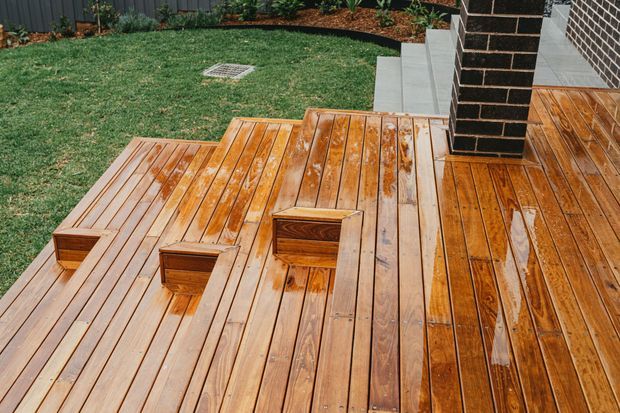 A polished timber outdoor deck with broad stepped platforms and integrated square wooden seating or planter boxes. Concrete steps lead around a dark brick column toward a landscaped lawn and garden bed, with a dark vertical fence in the background.
