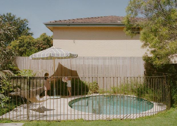 A suburban backyard with a small oval swimming pool enclosed by a metal safety fence. A striped parasol shades poolside seating, with timber fencing, a pale brick-clad house, tiled roof, and mature trees surrounding the space.