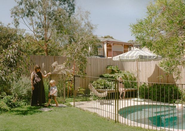 A landscaped residential backyard with a fenced swimming pool, curved pale paving, lawn, mature planting, and a large striped outdoor umbrella. A timber-clad or weatherboard house with a covered veranda is visible beyond the garden fence.