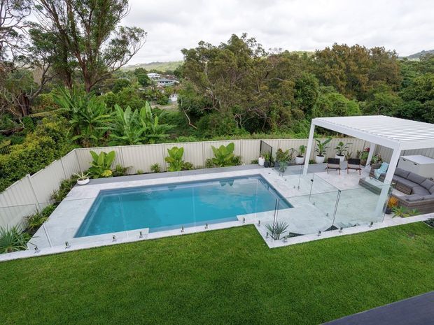 Elevated view of a landscaped backyard featuring a rectangular swimming pool, glass safety fencing, pale tiled paving, and a covered outdoor entertaining area with seating and an outdoor kitchen or bar. Dense greenery and mature trees surround the property, with a suburban hillside setting beyond.