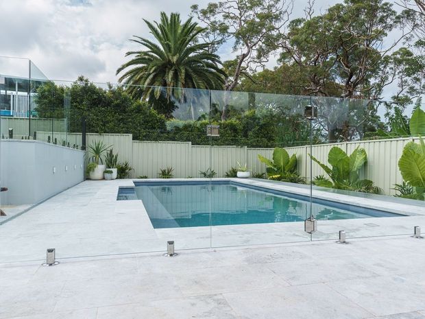 Modern residential backyard pool area with a rectangular swimming pool, pale stone or concrete paving, frameless glass safety fencing, and a light-coloured boundary fence. Tropical planting, including a large palm and broad-leaf plants, surrounds the pool, while part of a contemporary glazed house is visible at left.