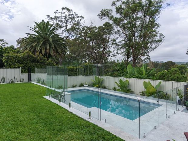 A landscaped backyard with a rectangular swimming pool enclosed by frameless glass safety fencing. The pool is surrounded by pale concrete paving and bordered by a white corrugated-style boundary fence, lush lawn, tropical planting and mature trees. The elevated outlook and overcast daylight create a calm, contemporary outdoor setting.