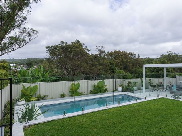 A landscaped backyard with a long, narrow in-ground swimming pool bordered by pale paving and frameless glass safety fencing. Tropical and native planting lines the perimeter, with a covered outdoor entertaining area and outdoor furniture at the far end. The property overlooks a lush, tree-filled suburban or semi-rural landscape beneath an overcast sky.