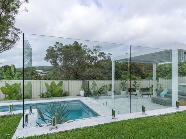 Contemporary outdoor pool area with a rectangular swimming pool, pale paved surrounds, frameless glass balustrade, and a white pergola-covered lounge zone. Tropical and subtropical planting lines a light corrugated boundary fence, with mature trees and neighboring homes visible beyond.