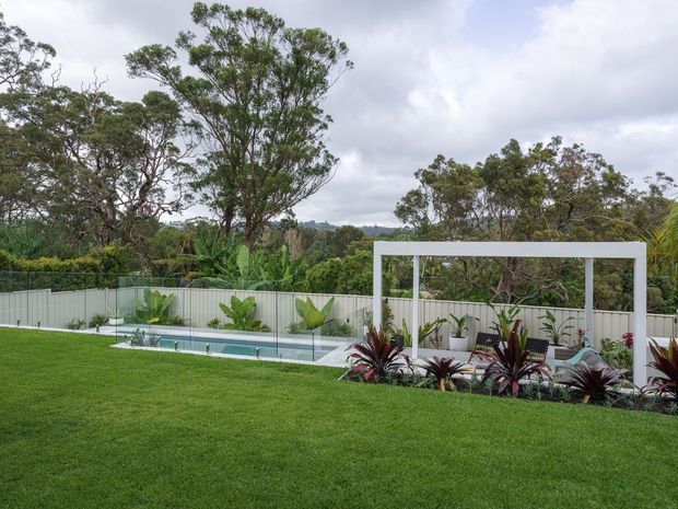 A landscaped backyard with a broad manicured lawn, a narrow in-ground swimming pool enclosed by glass fencing, tropical planting, and a simple white rectangular pergola or poolside structure. Mature trees and overcast sky form the background.