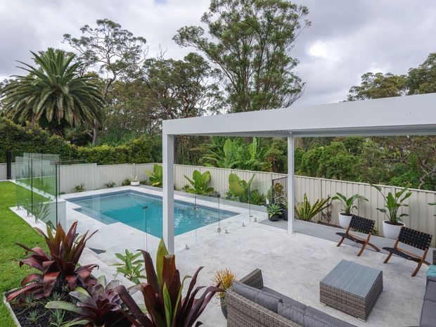 A contemporary outdoor entertaining area with a turquoise swimming pool, pale paved terrace, glass pool fencing, and a flat-roofed covered patio. The space includes an outdoor lounge setting and timber-framed chairs, with tropical planting and a white boundary fence creating a lush, private garden setting.