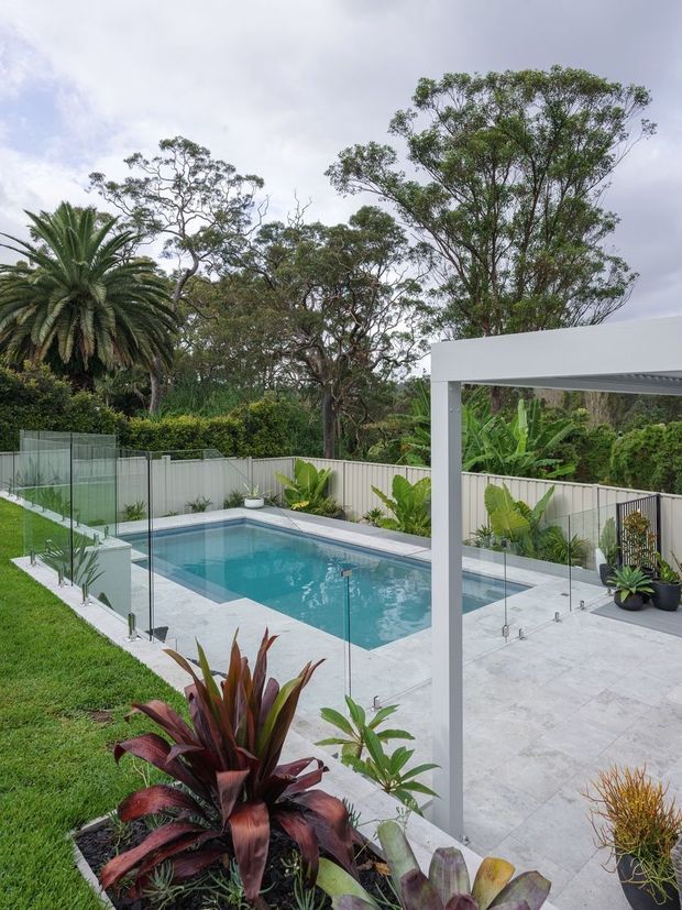 A landscaped backyard with a rectangular swimming pool enclosed by frameless glass safety fencing. Pale paved pool decking extends beneath a modern white flat-roofed outdoor structure, with lush tropical and subtropical planting, lawn, palms, and a light-coloured boundary fence.
