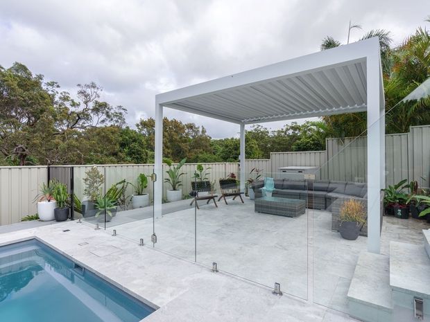 A contemporary outdoor entertaining area beside a swimming pool, featuring a white-framed louvred pergola, frameless glass pool fencing, pale stone paving, and an outdoor lounge with a sectional sofa and coffee table. Potted tropical and leafy plants line the perimeter, with a light grey boundary fence and dense greenery beyond.