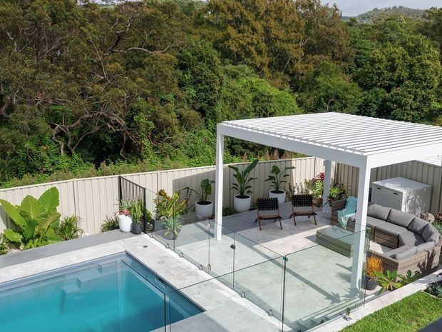 Elevated view of a landscaped outdoor entertaining area with a rectangular swimming pool, pale tiled paving, frameless glass pool fencing, and a white louvred pergola. The pergola shelters an outdoor lounge with a sectional sofa, dining chairs, potted plants, and a built-in outdoor setting, surrounded by dense greenery and a light privacy fence.