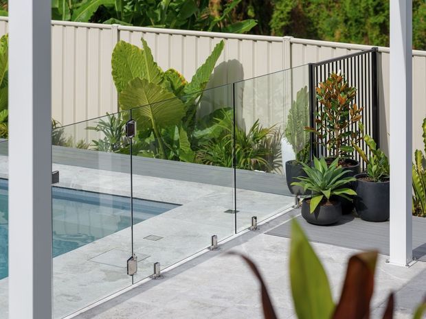 A contemporary outdoor pool area viewed from a covered terrace, featuring a pale tiled pool surround, frameless glass safety balustrade, white structural posts, a light vertical-panel boundary fence, and lush tropical planting in dark pots and garden beds.