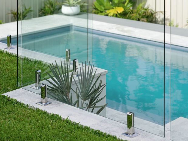 A contemporary outdoor swimming pool viewed through a frameless glass safety barrier. The pool has pale stone coping and surrounds, clear turquoise water, stainless-steel glass clamps or posts, and neatly landscaped tropical planting and lawn around it.