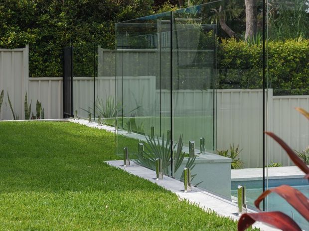 A landscaped outdoor pool area with a long, frameless glass pool fence and polished metal supports. The pool edge is visible at the lower right, bordered by pale paving and lush lawn, with a light privacy fence and dense planting behind.