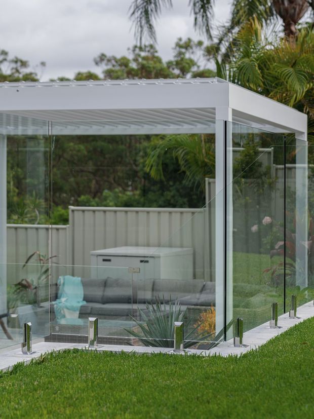 Exterior view of a modern glazed garden pavilion or outdoor room with a crisp white flat roof, extensive floor-to-ceiling glass walls, and frameless glass balustrading. A grey sofa is visible inside, with landscaped lawn and tropical planting surrounding the structure.