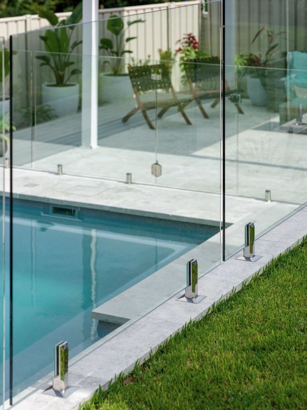 Close-up exterior view of a residential swimming pool bordered by pale stone paving and frameless glass safety balustrades with stainless steel fittings. The glass reflects a paved patio, potted tropical plants, outdoor seating, and a light privacy fence. A neatly maintained lawn borders the pool edge.
