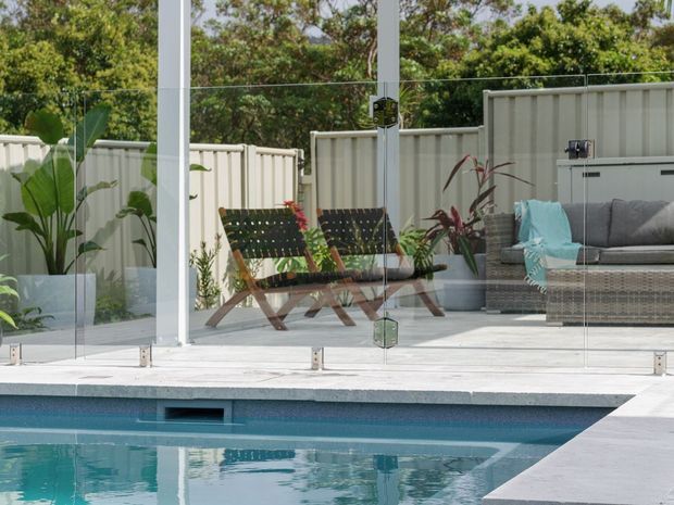 A landscaped outdoor pool area viewed through frameless glass pool fencing, with a pale tiled deck, white privacy fencing, tropical planting, and a compact outdoor lounge furnished with woven seating, cushions, and folding chairs.