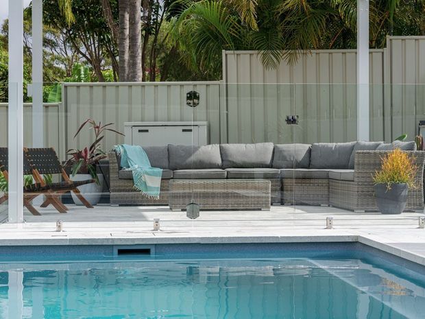 A bright outdoor pool area viewed through clear frameless glass fencing, with a turquoise swimming pool in the foreground and a covered outdoor lounge behind it. The lounge includes a large grey wicker sectional, woven armchairs, a low coffee table, soft blue throw, potted plants, and a pale vertical-panel privacy screen surrounded by tropical greenery.