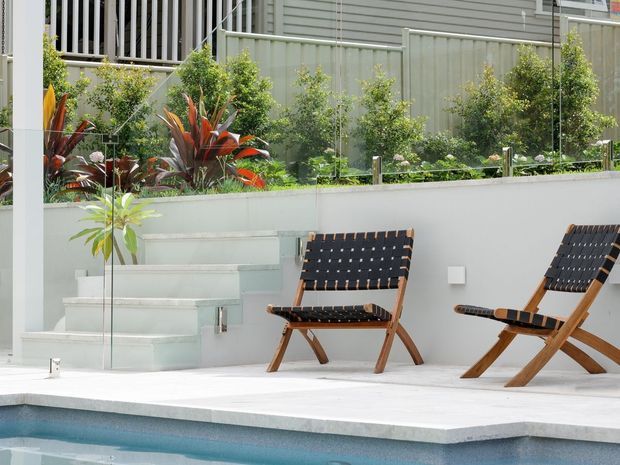 A contemporary outdoor pool area with a pale paved terrace, two timber-framed woven lounge chairs, a low white rendered retaining wall, glass balustrade, landscaped planting, and steps leading to an upper garden level. The edge of a rectangular swimming pool is visible in the foreground.