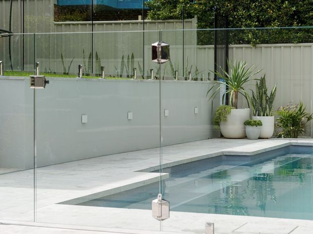 A modern outdoor swimming pool enclosed by frameless glass safety fencing, with pale stone paving, rendered boundary walls, and a selection of potted tropical and architectural plants. The scene has a clean, contemporary residential landscape design with calm neutral tones and turquoise pool water.