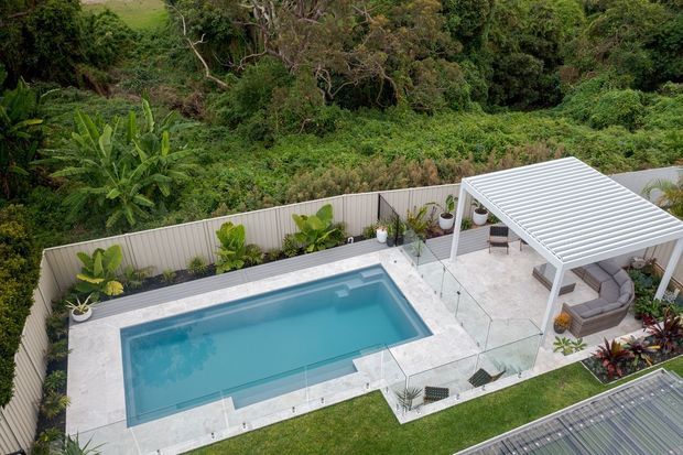 Elevated view of a landscaped residential pool area with a rectangular swimming pool, pale tiled or stone paving, glass pool fencing, planted borders, and a white slatted-roof outdoor pavilion containing an outdoor seating area. Dense tropical vegetation surrounds the property.