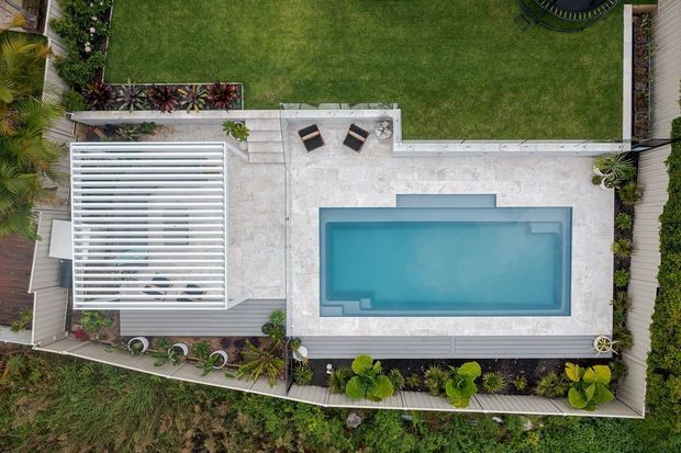 Aerial view of a landscaped residential backyard with a rectangular swimming pool, pale stone-paved pool terrace, outdoor lounge chairs, raised garden beds, lawn, and a large white louvred pergola or covered outdoor living structure.