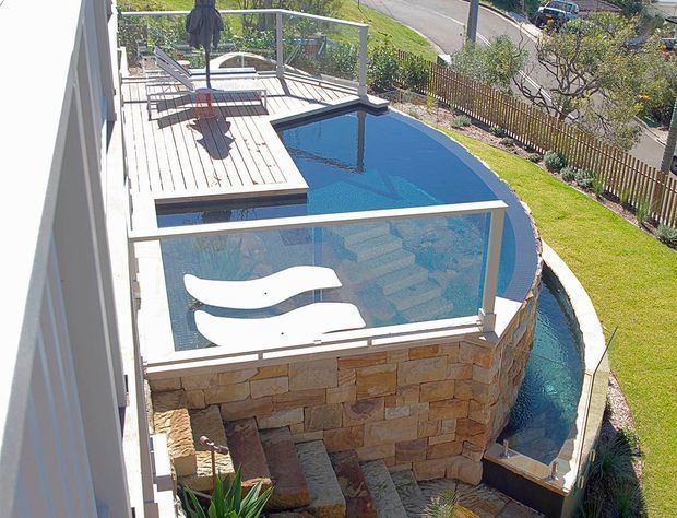Elevated view of a curved swimming pool beside a raised timber deck, enclosed by glass pool fencing and supported by a stacked natural-stone retaining wall. The pool includes sculptural white in-water loungers, with landscaped lawn and residential surroundings beyond.
