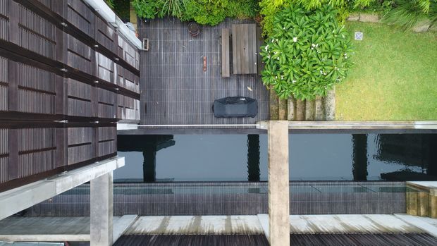 Aerial view of a narrow modern courtyard with a long rectangular reflecting pool or swimming pool bordered by dark timber decking. A dark-clad building and concrete structural elements frame the space, with dense tropical planting and a small lawn area along one edge.