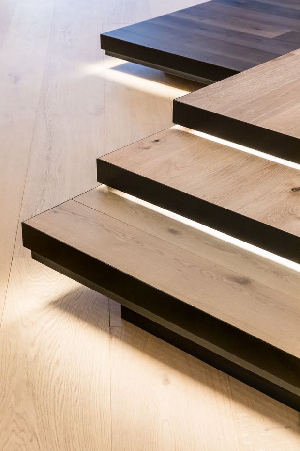 Close-up architectural detail of a floating timber staircase with broad light-oak treads, dark contrasting riser or support elements, and warm concealed LED lighting beneath each step. Matching timber flooring and wall panelling create a cohesive, refined interior finish.