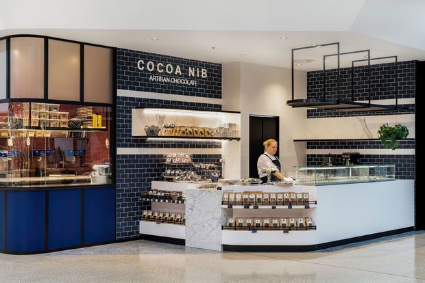 A modern artisanal chocolate shop interior with a central white stone sales counter, dark navy cabinetry, black subway-tile walls with white grout, illuminated product display shelving, and suspended black metal shelving above the counter. A curved glass-front display cabinet occupies the left side, while branded signage reads “COCOA NIB ARTISAN CHOCOLATE.” The space has a polished, refined retail fit-out with neutral terrazzo-like flooring and warm display lighting.