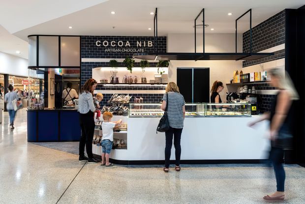 A contemporary artisan chocolate shop kiosk in a bright shopping-centre interior. The compact retail counter features a white and dark-blue base, glossy black subway-tile walls, illuminated display cases, open shelving with chocolate products, hanging black-framed glass display boxes, and prominent “COCOA NIB Artisan Chocolate” signage. Customers and staff occupy the foreground and counter area, while polished terrazzo flooring and neighbouring retail spaces are visible around the kiosk.