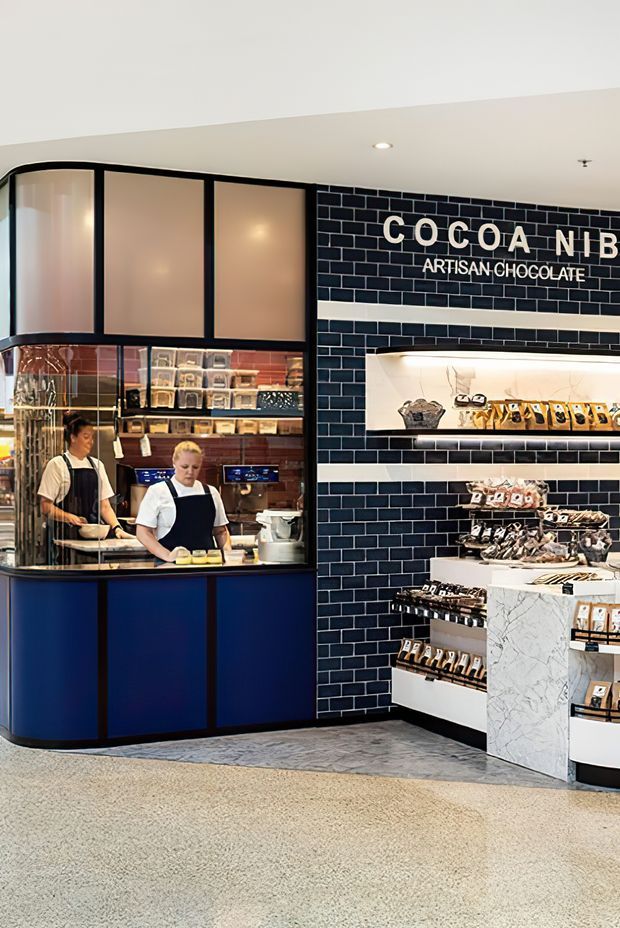 Interior view of a refined artisan chocolate shop, with a curved dark-blue and black service counter, large black-framed frosted glazing, navy blue subway-tile wall, illuminated recessed product shelving, marble-look display cabinetry, and terrazzo-style flooring. Staff and packaged chocolate products are visible behind and on the retail counters.