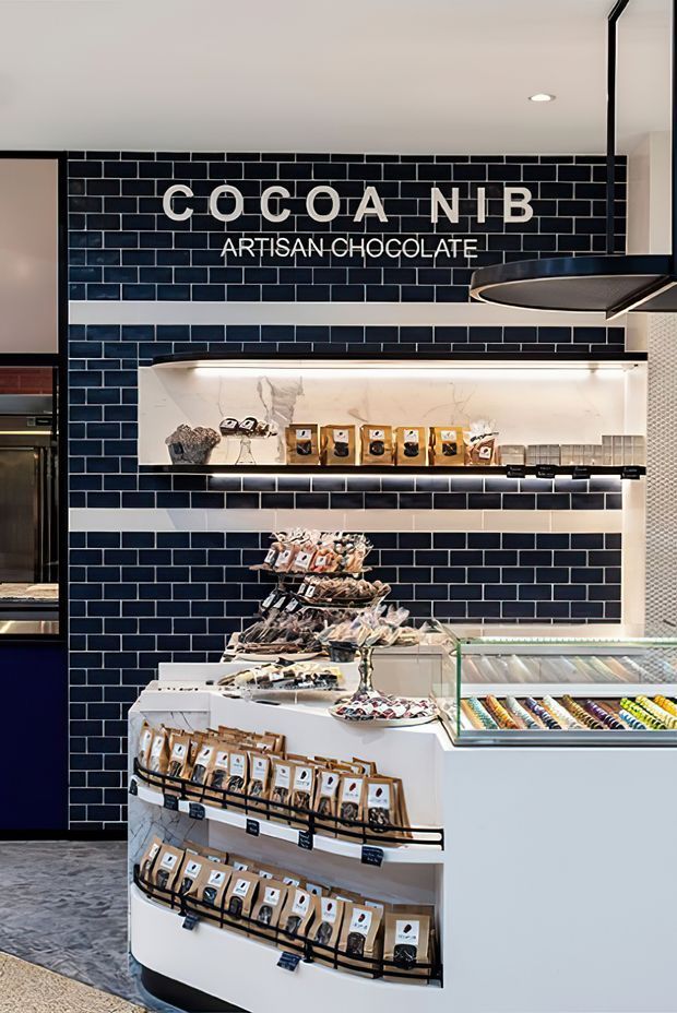 Interior view of a specialty artisan chocolate shop, dominated by a retail display wall clad in glossy navy-blue subway tiles with white grout. The wall features illuminated recessed shelving displaying packaged chocolate products, while a curved white freestanding display unit and glass-topped refrigerated confectionery case occupy the foreground. A black suspended range-style canopy or feature fixture is visible overhead, and polished grey flooring extends through the space.