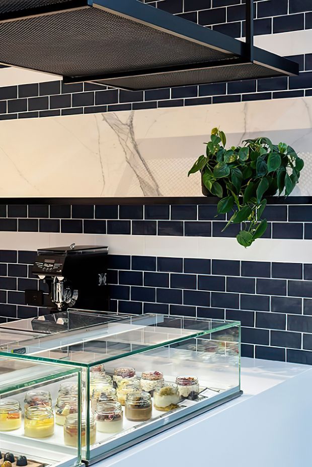 A stylish café or dessert shop interior featuring a black subway-tile wall with contrasting white grout and a large marble-look feature panel. A black espresso machine sits behind a glass display counter filled with individual desserts in jars. Dark metal mesh shelving and a trailing green plant add industrial character, while the white counter surfaces keep the space bright and crisp.