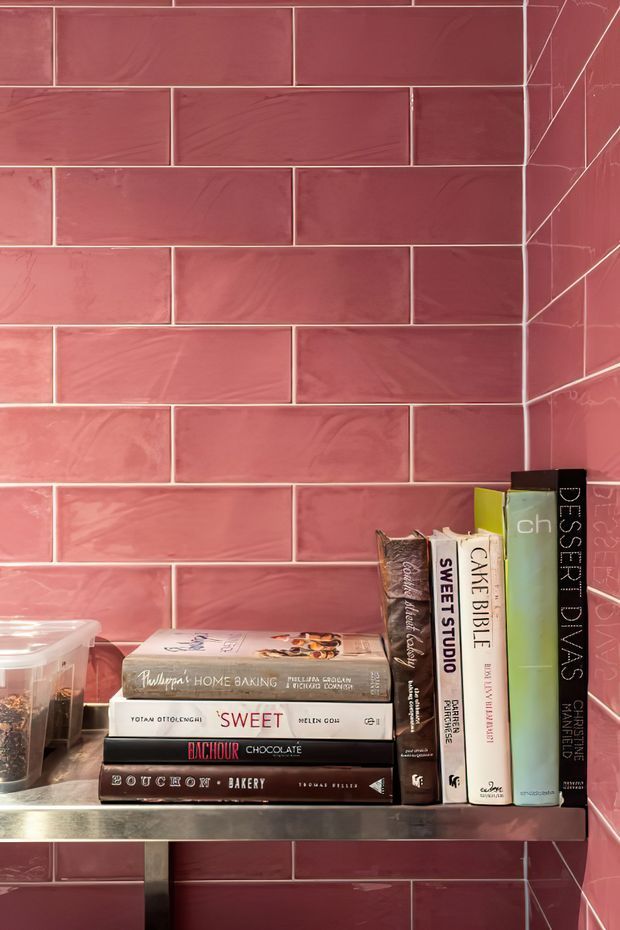 Close-up of a built-in shelf niche lined with glossy pink-red rectangular subway tiles in a running-bond pattern. The niche contains neatly arranged baking and dessert cookbooks, with a metal shelf and dark vertical support visible at the bottom. The image is brightly lit and tightly framed around the tiled joinery and books.