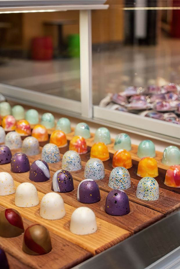 Close-up of a display counter in a confectionery or food retail space, with rows of colourful dome-shaped sweets arranged on wooden trays. A large glass display case and reflected merchandise are visible in the background. The image is tightly framed on the product display rather than the surrounding room.