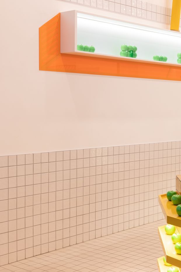 A brightly lit tiled commercial interior with pale pink square tiles covering the lower walls and floor. A vivid orange built-in wall display shelf with white illuminated niches runs across the upper wall, holding green apple-like display objects. Matching orange shelving with green produce is partially visible at the right edge. The space has a playful, highly saturated retail or showroom aesthetic.