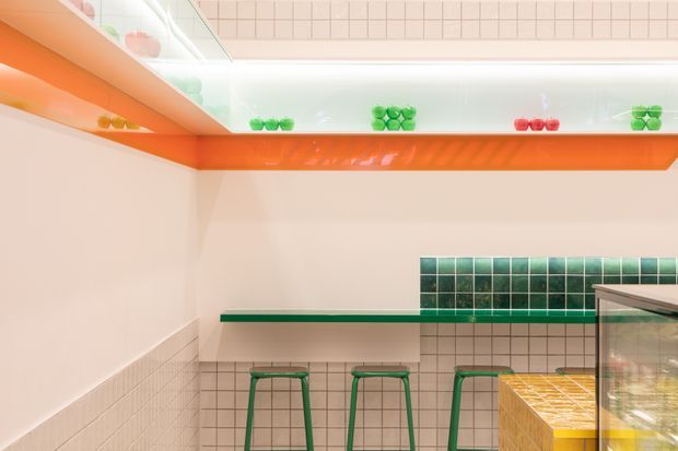 Bright, playful café or food-service interior with pale tiled walls, vivid orange illuminated shelving and trim, green tiled backsplash, matching green counter ledge and stools, and a tiled service counter with glass display case. The space has a clean contemporary fit-out with strong retro-inspired colour accents.