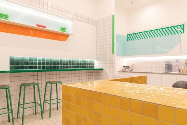 Bright contemporary kitchen or food-preparation space featuring white square wall tiles, a large yellow-orange tiled island or counter, green tiled shelving and green metal stools. A wall-mounted green rack, open shelving, pale cabinetry, and under-shelf lighting create a playful, graphic interior with strong green and orange accents.