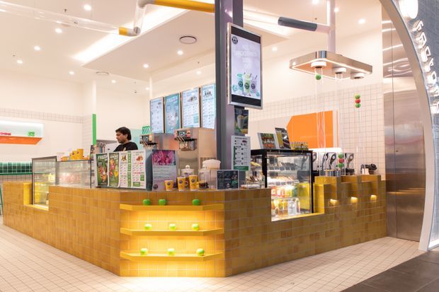 Bright modern commercial food-service counter with yellow tiled cabinetry, illuminated display shelving, refrigerated glass cases, menu boards, and stainless-steel equipment. The compact retail interior uses a white tiled and plastered fit-out with strong yellow and orange accents.