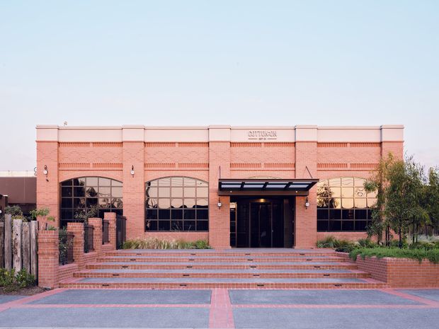 Front elevation of a single-storey commercial brick building with a symmetrical facade, large arched black-framed windows, a central glazed entry beneath a dark metal canopy, broad brick steps, and landscaped planting. The building has decorative brick detailing and a warm terracotta-red brick exterior.