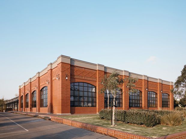 Exterior view of a substantial single-storey red-brick building with a long, articulated facade, tall black-framed arched industrial-style windows, decorative brick detailing, and a parapet roofline. The building fronts a paved parking area and landscaped verge with low hedges and ornamental grasses.
