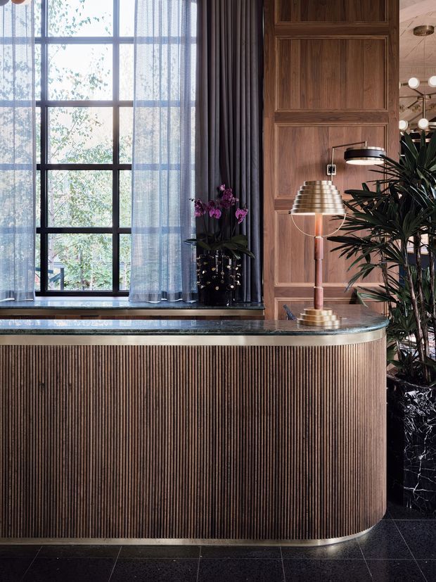A refined reception or bar-style joinery area featuring a curved fluted timber-clad counter with a dark stone benchtop, richly panelled timber wall, black-framed multi-pane window with sheer and dark curtains, and brass-toned decorative lamps. A tall indoor plant and floral arrangement add natural contrast. The overall scheme is dark, warm, and luxurious with Art Deco-inspired detailing.