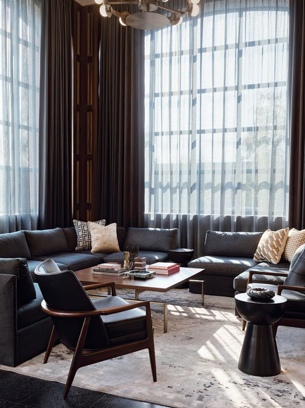 A sophisticated living room with a large dark sectional sofa arranged around a low rectangular coffee table, accompanied by mid-century-inspired lounge chairs and sculptural side tables. Floor-to-ceiling windows with sheer patterned blinds and heavy dark curtains create strong natural light and shadows across a pale area rug. The room features dark timber wall panelling, a sculptural branching pendant light, and a refined, moody palette of charcoal, brown, cream, and warm wood.
