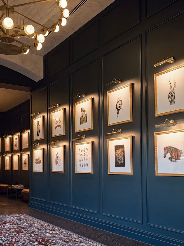 A sophisticated, moody interior gallery or hospitality space with deep navy-blue paneled walls displaying numerous gold-framed artworks. Brass picture lights illuminate each frame, while an ornate contemporary chandelier with warm globe bulbs hangs from a decorative ceiling. A patterned area rug and dark flooring add richness to the corridor-like space.