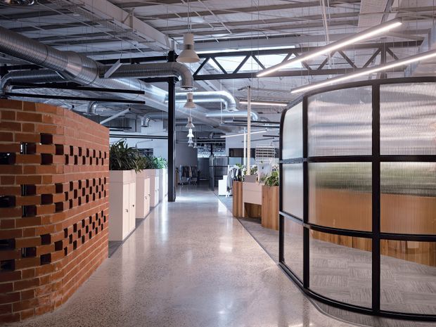 A spacious contemporary commercial office interior with an open-plan layout. The corridor features polished concrete or terrazzo flooring, exposed services and ductwork, suspended linear lighting, a perforated red-brick feature wall, timber-toned cabinetry and planters, and curved black-framed translucent glazed meeting-room partitions. The high ceiling and industrial detailing create a modern adaptive workplace atmosphere.