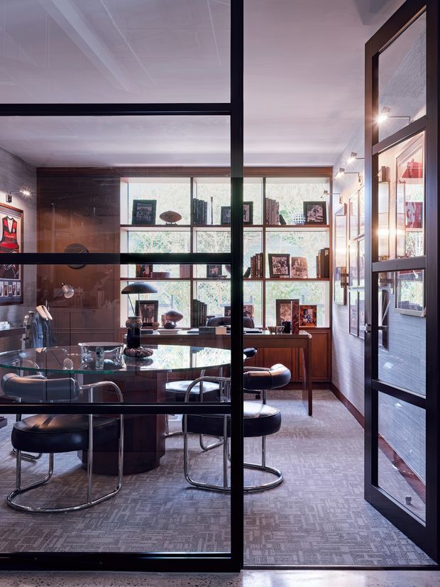 A refined home office viewed through black-framed glazed doors, with a dark timber built-in wall and shelving, large windows, a polished desk, black leather cantilever chairs, artwork, books, and patterned carpet. The room has a sophisticated, richly layered palette with strong natural light and interior glazing.