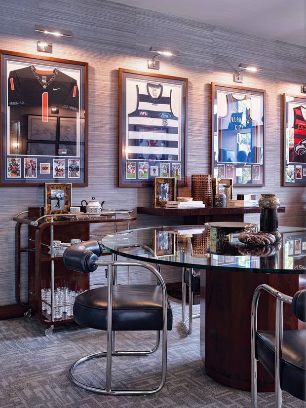 A sophisticated sports memorabilia lounge or home bar featuring framed sports jerseys and collectible displays on a textured grey wall. The room includes a glossy dark wood bar or dining counter, glass-topped console tables, black leather tubular-metal chairs, polished metal accents, display shelving, and patterned carpet. Integrated picture lights illuminate the framed jerseys, creating a refined, masculine atmosphere.