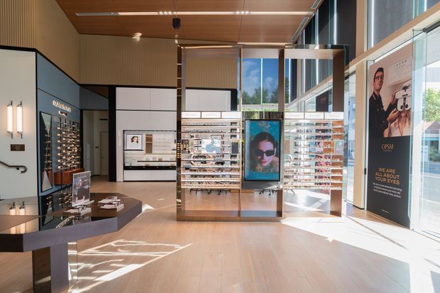 A bright contemporary optical retail showroom with floor-to-ceiling glazing, warm timber ceilings and flooring, integrated display shelving, freestanding product racks, framed eyewear displays, and branded promotional graphics. The space has a clean, refined commercial fit-out with abundant natural light.