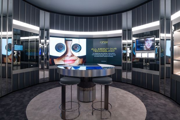 A dark, highly polished commercial optical retail or consultation space with a curved circular layout. Mirrored and vertically panelled walls surround a central round metallic display or consultation table with stools. Integrated illuminated display screens and large branded eye-care graphics form a feature wall, while concealed linear lighting creates a sleek, futuristic atmosphere.